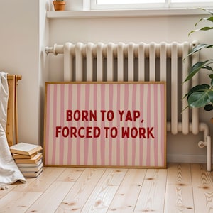 May include: A framed print with a pink and white striped design and the red text "BORN TO YAP, FORCED TO WORK". The artwork is displayed in a room with a radiator, a plant in a terracotta pot, and a stack of books.
