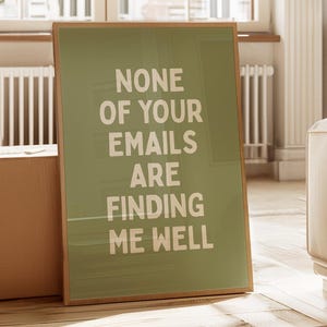 May include: A rectangular framed wall art piece featuring a muted olive green background with the white text "NONE OF YOUR EMAILS ARE FINDING ME WELL" printed in a bold, sans-serif font. The frame is a light wood tone with a clean, simple border.