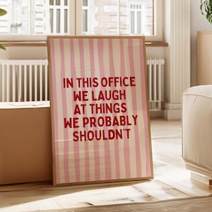 Może przedstawiać: Oprawiony wydruk z różowo-białym paskowanym tłem. Wydruk zawiera czerwony tekst "IN THIS OFFICE WE LAUGH AT THINGS WE PROBABLY SHOULDN'T". Rama jest w kolorze jasnobrązowym.