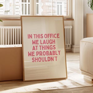 Op de afbeelding: Een ingelijste print met de tekst "IN THIS OFFICE WE LAUGH AT THINGS WE PROBABLY SHOULDN'T" in roze letters. De print zit in een lichtgekleurde houten lijst en leunt tegen een muur in een kamer met een raam.