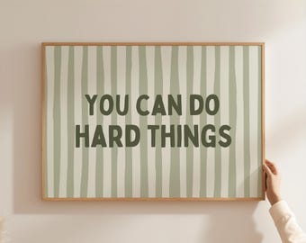 Op de afbeelding: Een ingelijste print met de tekst "YOU CAN DO HARD THINGS" in donkergroene letters. De achtergrond heeft verticale strepen in lichtgroen en wit. Het frame is gemaakt van lichtbruin hout.