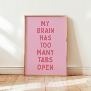 Op de afbeelding: Een ingelijste roze kunstprint met de tekst "MY BRAIN HAS TOO MANY TABS OPEN" in een donkerdere roze lettertype. Het kunstwerk wordt tentoongesteld op een houten vloer tegen een witte muur.