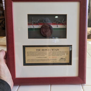 Może przedstawiać: Oprawiony artefakt historyczny z drutem kolczastym, pieczęcią woskową i certyfikatem. Rama jest w kolorze ciemnoczerwonym. Na certyfikacie widnieje napis "THE IRON CURTAIN" i "Certificate of origin". Drut kolczasty umieszczono na tle zielono-biało-czerwonych pasków.