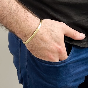 May include: A gold-colored, minimalist cuff bracelet. The bracelet is a simple, solid band with a smooth, matte finish. The bracelet is worn on a wrist, with the person wearing a black shirt and blue jeans.