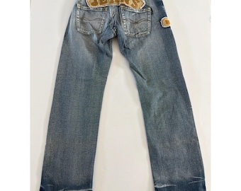 Puede incluir: Vaqueros de mezclilla azul claro con un parche de tela beige sobre los bolsillos traseros con la palabra JEANS bordada en marrón. El pantalón incluye una etiqueta de cuero sintético marrón en la cintura, dos bolsillos traseros con costuras y dobladillos deshilachados.