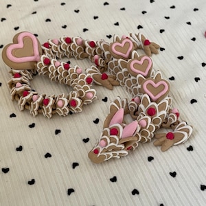May include: A brown and white dragon figurine with pink heart accents and red dot details. The dragon is made of multiple segments and is posed on a white fabric with black heart shapes.