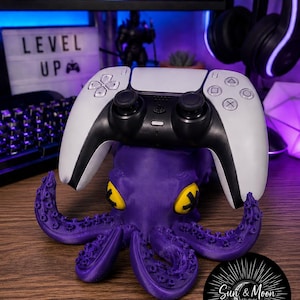 May include: A purple octopus-shaped controller stand with yellow eyes and black pupils, featuring textured tentacles. A white and black wireless game controller rests on top of the octopus figure, which is designed to hold gaming peripherals.