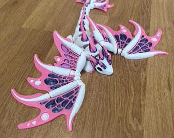 May include: A white and pink dragon figurine with intricate wing details. The dragon has a long neck and tail, with pink accents on the wings and spines. The wings have a dark blue webbed pattern. The figurine is posed on a wooden surface.