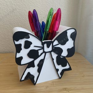 May include: A white pen holder with a black and white cow print bow design. The holder contains several colorful pens in red, purple, blue, green, and pink. The pen holder is on a wooden surface.