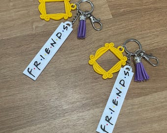 May include: Two keychains featuring a yellow picture frame design and the word "FRIENDS" in black lettering on a white background. Each keychain has a silver keyring, a purple tassel, and a clasp.