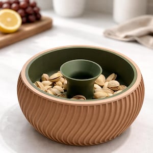 May include: A two-tone nut bowl with a ridged exterior. The bowl has a light brown exterior and a dark green interior. The center of the bowl has a dark green cup. The bowl is filled with pistachios.
