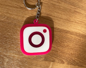 May include: A pink and white Instagram logo keychain with a silver key ring and chain. The keychain features the iconic camera logo with a white background, a pink border, and a dark pink circle and dot.