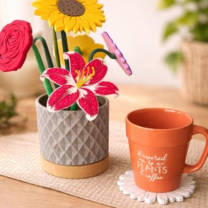 May include: A decorative arrangement of colorful artificial flowers in a gray patterned pot with a cork base. An orange mug with the text "Powered by Plants & Coffee" sits on a white daisy-shaped coaster. The image is on a neutral-toned surface.