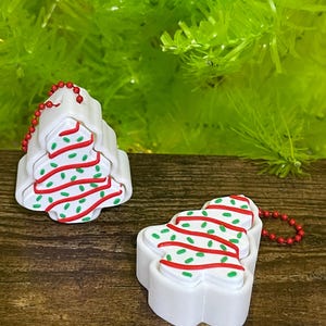 May include: Two white Christmas tree-shaped ornaments with red and green detailing. Each ornament has a red beaded chain. The ornaments are on a wooden surface with a green background.