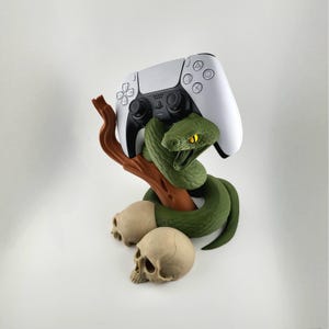 May include: A white PlayStation 5 controller rests on a brown branch, held by a green snake with yellow eyes. Two skulls are at the base. The controller is a gaming accessory.