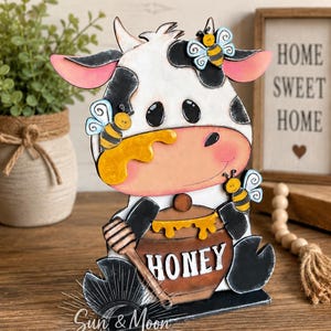 May include: A decorative wooden cutout of a white cow with black spots, featuring three small bees, a honey pot labeled HONEY, and a honey dipper. The cow has pink ears and a peach-colored snout. The base includes the text Sun & Moon STUDIOS.