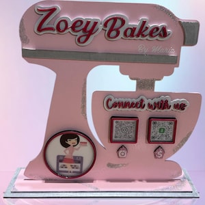 May include: Pink stand-up cutout of a kitchen mixer with the words "Zoey Bakes" and "By Maria" on the top. The mixer has a white bowl and a red rim. There are two QR codes on the front of the mixer with the words "Connect with us".