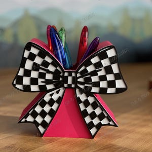 May include: A pink pen holder shaped like a gift box with a large bow. The bow has a black and white checkered pattern. Several colorful pens are inside the holder. The background is a blurred image of a landscape.