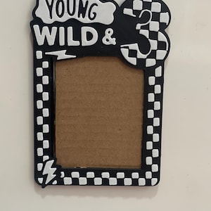 May include: Black and white photo frame with the text "YOUNG WILD &" and the number "3". The frame has a checkered pattern and a lightning bolt design. The frame is designed to hold a photo.