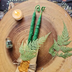 以下が含まれることがあります： 茶色の紙に包まれた緑色のろうそく2本。金色のワックスシールと緑色のシダの葉が飾られています。ろうそくは、小さなろうそくとガラス瓶のある木製の表面の上に置かれています。