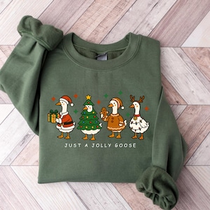 May include: Green crewneck sweatshirt featuring a festive design with four cartoon geese dressed in Christmas attire. The geese are wearing Santa hats, a Christmas tree costume, a gingerbread outfit, and reindeer antlers. The text "JUST A JOLLY GOOSE" is printed below the geese.