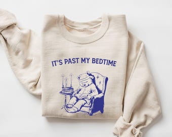 Può includere: Felpa color crema con la scritta blu "IT'S PAST MY BEDTIME". La grafica raffigura un orso con la cuffia da notte che legge in una poltrona blu. Un tavolino con candele e libri è accanto all'orso. Le maniche sono arrotolate.