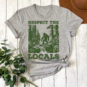 May include: Gray t-shirt with a green graphic of a Bigfoot walking through a forest with the text "Respect the Locals".