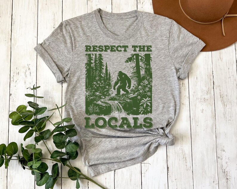 May include: Gray t-shirt with a green graphic of a Bigfoot walking through a forest with the text "Respect the Locals".