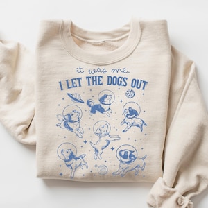 May include: Beige sweatshirt with a blue graphic of dogs in space helmets and planets. The text "it was me. I LET THE DOGS OUT" is printed above the graphic. The sweatshirt has long sleeves and a crew neck.