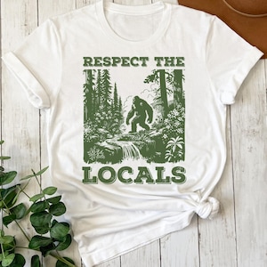 May include: White t-shirt with a green graphic print of a Bigfoot walking through a forest. The text "Respect the Locals" is printed below the image.