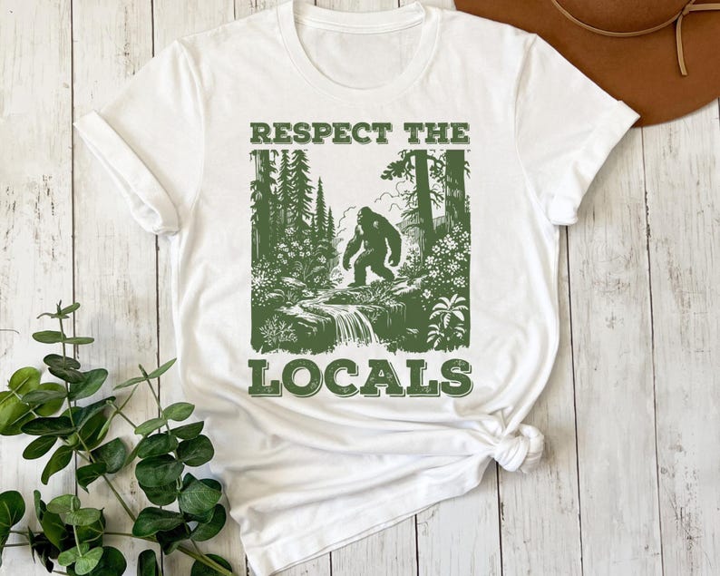 May include: White t-shirt with a green graphic print of a Bigfoot walking through a forest. The text "Respect the Locals" is printed below the image.