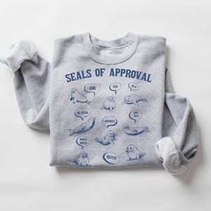 Peut inclure: Sweat-shirt gris avec un graphique de phoques avec des bulles de dialogue qui disent "OK", "Yep", "N-uh", "Oh yeah", "I Approve", "Dope That", "Roger Roger", "Defo". Le texte "Seals of Approval" est en haut du graphique.