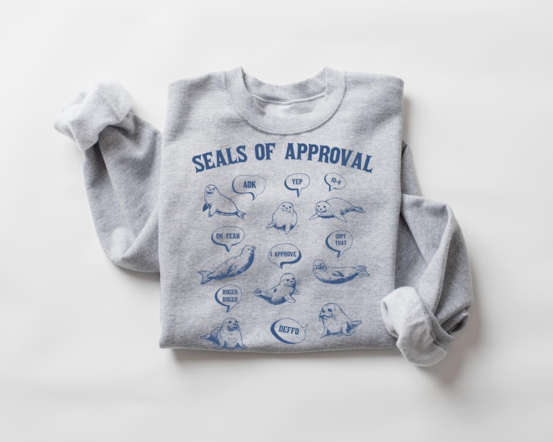 Peut inclure: Sweat-shirt gris avec un graphique de phoques avec des bulles de dialogue qui disent "OK", "Yep", "N-uh", "Oh yeah", "I Approve", "Dope That", "Roger Roger", "Defo". Le texte "Seals of Approval" est en haut du graphique.