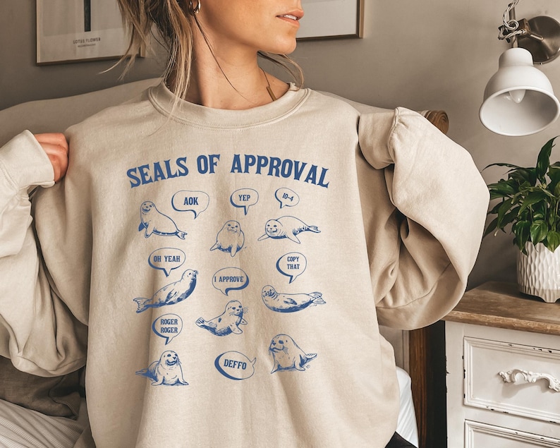 Peut inclure: Un sweat-shirt beige avec le texte "Seals of Approval" imprim&eacute; sur le devant. Le texte est entour&eacute; d'illustrations de phoques avec diff&eacute;rentes bulles de dialogue, chacune contenant une phrase diff&eacute;rente. Les phrases incluent "AOK", "Yep", "B-4", "Oh Yeah", "I Approve", "Copy That", "Roger Roger", "Deffo".