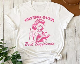 May include: White t-shirt with pink text that reads "Crying over Book Boyfriends". A cartoon illustration of a woman with tears in her eyes holding a book and a cup of coffee is featured on the shirt.