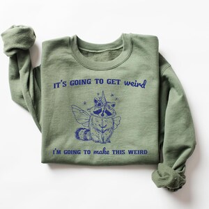May include: Green sweatshirt with a blue graphic of a raccoon wearing a unicorn horn and fairy wings. The text "It's going to get weird" and "I'm going to make this weird" is printed on the sweatshirt.
