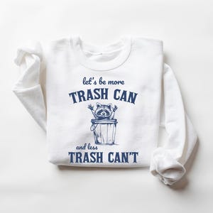 Pu&ograve; includere: Maglietta bianca a maniche lunghe con testo blu che recita "let's be more trash can and less trash can't". Un procione dei cartoni animati &egrave; dentro un bidone della spazzatura.