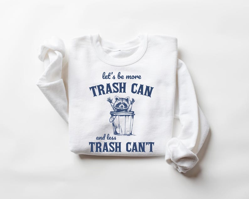 Pu&ograve; includere: Maglietta bianca a maniche lunghe con testo blu che recita "let's be more trash can and less trash can't". Un procione dei cartoni animati &egrave; dentro un bidone della spazzatura.