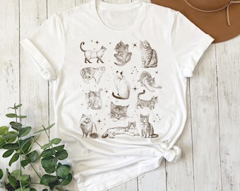 May include: White t-shirt with a print of various cats in different poses. The cats are all in shades of brown and gray, and the print is surrounded by small stars.