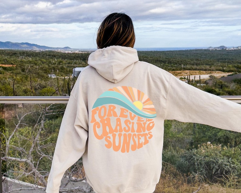 May include: A cream-colored hoodie with a graphic on the back that says "FOREVER CHASING SUNSETS" in a retro font with a colorful sunset and wave design.