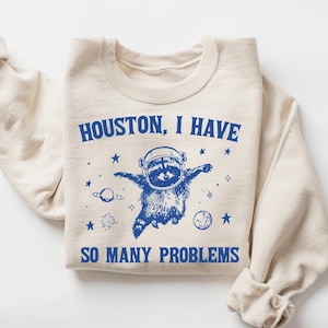 May include: Cream-colored sweatshirt with blue graphic of a raccoon in a space helmet, surrounded by stars and planets. Text reads "Houston, I have so many problems."