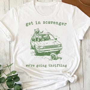 Op de afbeelding: Wit t-shirt met een groene afbeelding van wasberen in een auto. De tekst "get in scavenger" staat boven de auto en "we're going thrifting" eronder. Het ontwerp is een speelse illustratie.