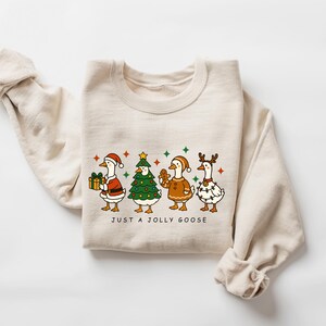 May include: A beige sweatshirt featuring a festive graphic of four cartoon geese dressed in holiday attire. One goose holds a gift, another is a Christmas tree, and others wear festive lights and reindeer antlers. The text "JUST A JOLLY GOOSE" is below the geese.
