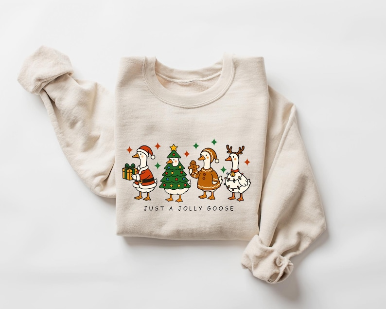 May include: A beige sweatshirt featuring a festive graphic of four cartoon geese dressed in holiday attire. One goose holds a gift, another is a Christmas tree, and others wear festive lights and reindeer antlers. The text "JUST A JOLLY GOOSE" is below the geese.