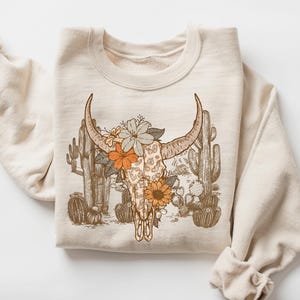 May include: A beige sweatshirt with a graphic print of a cow skull with floral accents and cacti. The skull is surrounded by flowers in shades of orange, yellow, and pink.