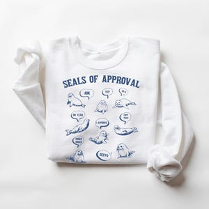 Peut inclure: Sweat-shirt blanc avec un graphique bleu de phoques avec des bulles de dialogue qui disent "AOK", "Yep", "Oh Yeah", "J'approuve", "Copie &ccedil;a", "Deffo", et "Plus grand, plus grand". Le texte "Seals of Approval" est en haut du graphique.