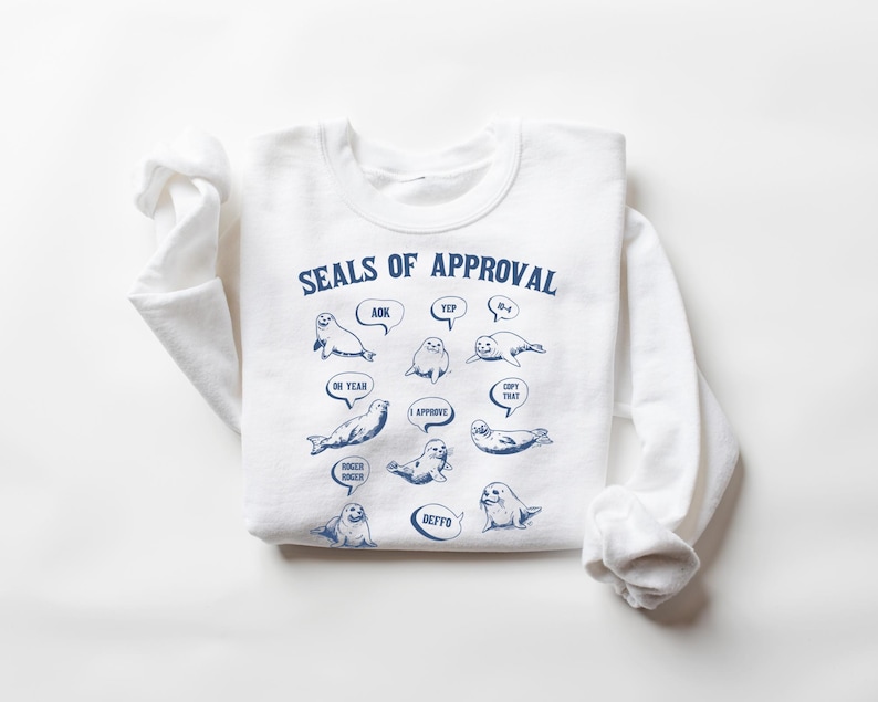 Peut inclure: Sweat-shirt blanc avec un graphique bleu de phoques avec des bulles de dialogue qui disent "AOK", "Yep", "Oh Yeah", "J'approuve", "Copie &ccedil;a", "Deffo", et "Plus grand, plus grand". Le texte "Seals of Approval" est en haut du graphique.