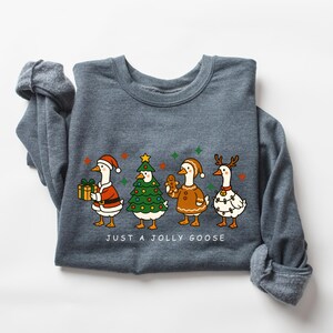 May include: A dark gray sweatshirt featuring a festive graphic of four geese dressed in holiday attire. One goose holds a gift, another is a Christmas tree, a third is a gingerbread cookie, and the last is decorated with lights. The text "JUST A JOLLY GOOSE" is below.