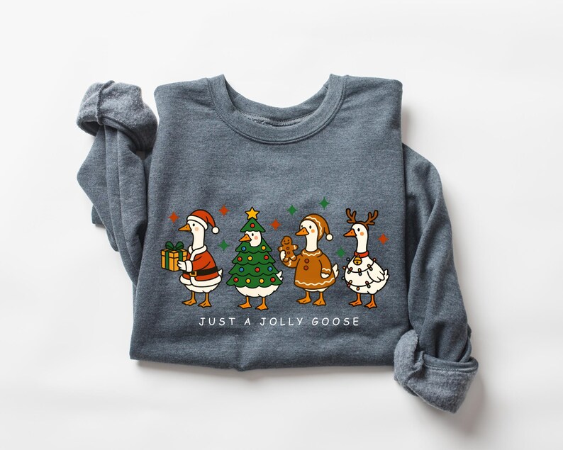 May include: A dark gray sweatshirt featuring a festive graphic of four geese dressed in holiday attire. One goose holds a gift, another is a Christmas tree, a third is a gingerbread cookie, and the last is decorated with lights. The text "JUST A JOLLY GOOSE" is below.