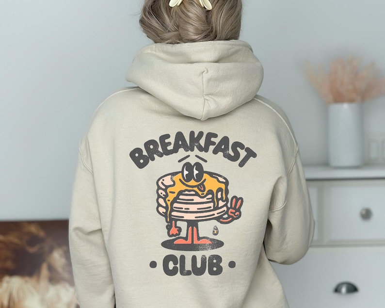 May include: A beige hoodie with a cartoon illustration of a pancake stack with a face and the text "BREAKFAST CLUB".